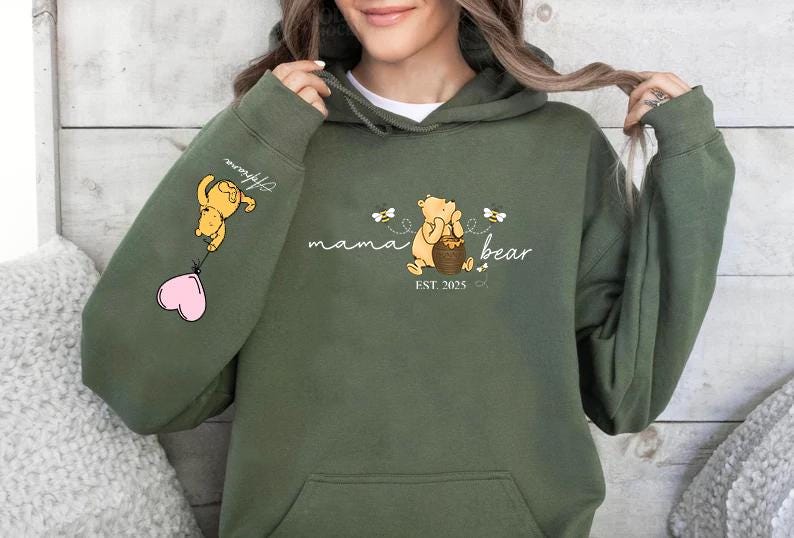 May include: Green hooded sweatshirt with a Winnie the Pooh inspired design. The text reads "mama bear est. 2023" with a bear holding a honey pot and bees flying around. The left sleeve has a Winnie the Pooh character holding a pink heart balloon.
