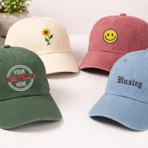 May include: Four baseball caps in different colours: green with "YOUR DESIGN HERE" text, cream with a sunflower, red with a smiley face, and light blue with "Huxley" text. The caps have curved peaks and adjustable straps.