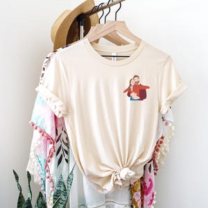 Pu&ograve; includere: T-shirt color crema con una piccola grafica di una coppia che si abbraccia. La maglietta &egrave; annodata in vita e appesa a una gruccia di legno. Altri capi di abbigliamento, tra cui un cappello di paglia e uno scialle fantasia, sono anche sull'appendiabiti.