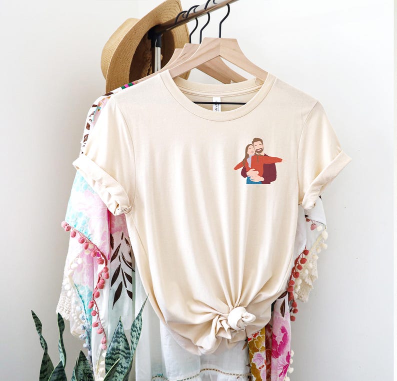 Pu&ograve; includere: T-shirt color crema con una piccola grafica di una coppia che si abbraccia. La maglietta &egrave; annodata in vita e appesa a una gruccia di legno. Altri capi di abbigliamento, tra cui un cappello di paglia e uno scialle fantasia, sono anche sull'appendiabiti.