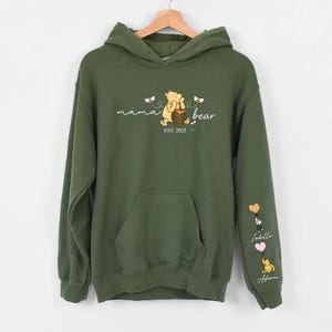 May include: A green hooded sweatshirt with a Winnie the Pooh character and the text "mama bear est. 2023". The sleeves have embroidered hearts with names.