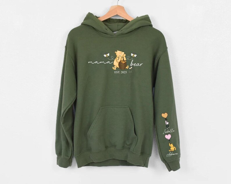 May include: A green hooded sweatshirt with a Winnie the Pooh character and the text "mama bear est. 2023". The sleeves have embroidered hearts with names.