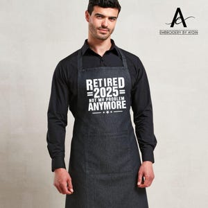 Puede incluir: Delantal gris oscuro con un bolsillo frontal grande y el texto blanco "RETIRED 2025 = NOT MY PROBLEM ANYMORE". El delantal lo lleva una persona con una camisa negra.