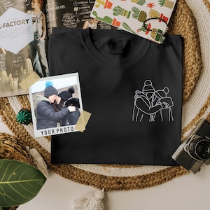 May include: Black t-shirt featuring a white outline of three people embracing. The t-shirt has a white Polaroid frame with a photo of two people hugging in front of a snowy background. The text "YOUR PHOTO" is printed below the photo.