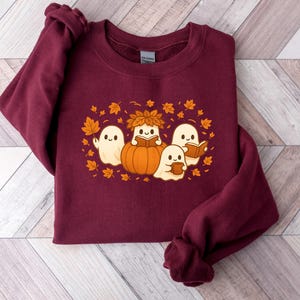 Peut inclure: Un sweat-shirt bordeaux avec un motif sur le th&egrave;me de l'automne. Le motif repr&eacute;sente quatre fant&ocirc;mes de dessins anim&eacute;s, une citrouille et des feuilles d'automne. Deux fant&ocirc;mes lisent des livres, un tient une tasse et l'autre une feuille. Le motif est dans les tons orange, blanc et marron.