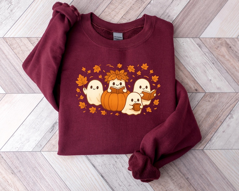 Peut inclure: Un sweat-shirt bordeaux avec un motif sur le th&egrave;me de l'automne. Le motif repr&eacute;sente quatre fant&ocirc;mes de dessins anim&eacute;s, une citrouille et des feuilles d'automne. Deux fant&ocirc;mes lisent des livres, un tient une tasse et l'autre une feuille. Le motif est dans les tons orange, blanc et marron.