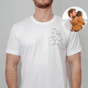 以下が含まれることがあります： 子供を抱いている人の黒い線画が描かれた白いTシャツ。小さな円形の画像には、子供を抱いている人が写っています。Tシャツは柔らかい素材でできており、半袖です。