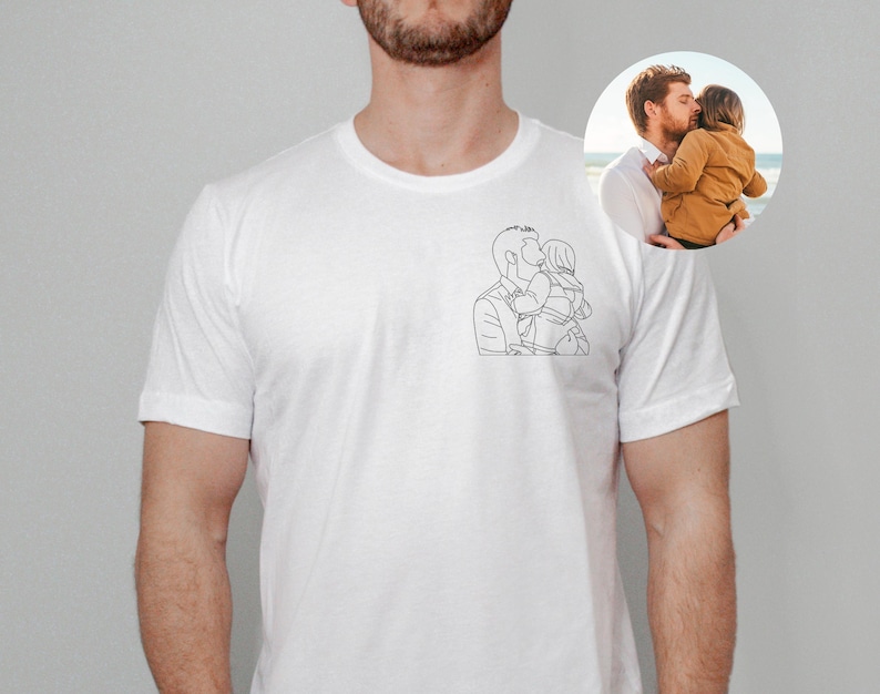 以下が含まれることがあります： 子供を抱いている人の黒い線画が描かれた白いTシャツ。小さな円形の画像には、子供を抱いている人が写っています。Tシャツは柔らかい素材でできており、半袖です。
