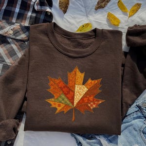 May include: A brown sweatshirt featuring a large, colorful maple leaf design. The leaf is composed of various patchwork patterns in shades of orange, red, green, and cream. The sweatshirt is folded on a white surface with a plaid shirt and jeans.