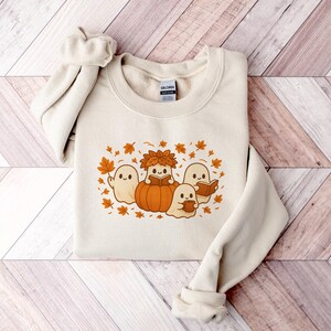 Peut inclure: Sweat-shirt cr&egrave;me avec un motif sur le th&egrave;me de l'automne. Le motif comprend des fant&ocirc;mes de dessins anim&eacute;s, une citrouille et des feuilles d'automne dans des tons orange et marron. Le sweat-shirt a un col rond et des manches longues.