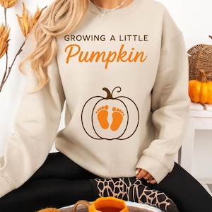 May include: A beige sweatshirt featuring the text "GROWING A LITTLE Pumpkin" in orange. A brown pumpkin graphic with orange baby footprints is centered on the front. The sweatshirt is being worn.