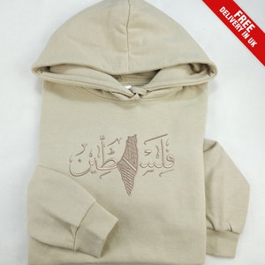 Op de afbeelding: Een beige hoodie met een grafische print van de Palestijnse vlag en Arabische tekst. De tekst luidt "فلسطين", wat "Palestina" betekent.