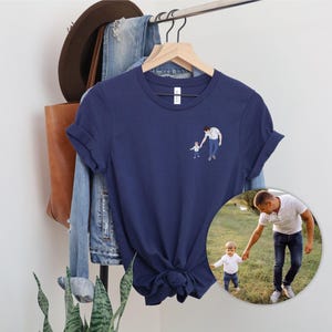 Pu&ograve; includere: T-shirt blu navy con una piccola illustrazione di un genitore e un bambino che si tengono per mano. La maglietta &egrave; appesa a un gancio di legno con altri vestiti e accessori sullo sfondo. Una foto circolare della stessa coppia &egrave; visibile in basso a destra.