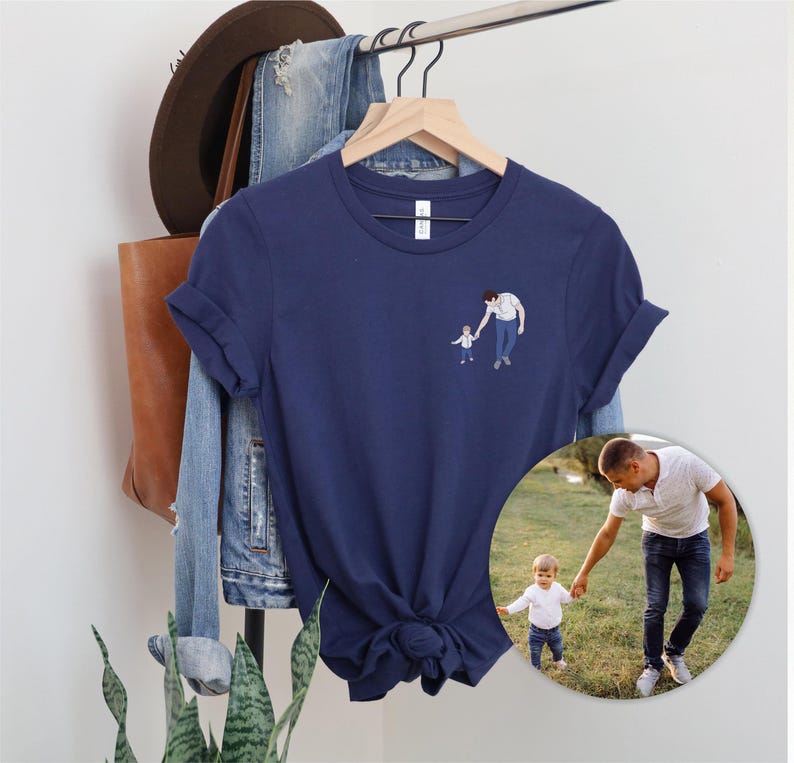 Pu&ograve; includere: T-shirt blu navy con una piccola illustrazione di un genitore e un bambino che si tengono per mano. La maglietta &egrave; appesa a un gancio di legno con altri vestiti e accessori sullo sfondo. Una foto circolare della stessa coppia &egrave; visibile in basso a destra.
