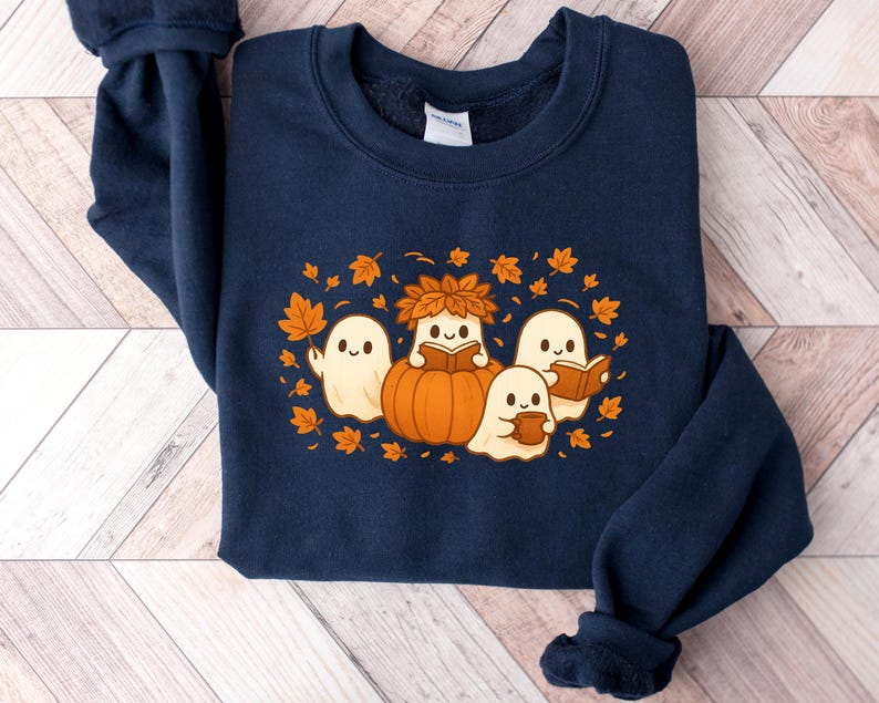 Peut inclure: Sweat-shirt bleu marine avec un motif sur le th&egrave;me de l'automne. Le motif comprend des fant&ocirc;mes de dessins anim&eacute;s, une citrouille et des feuilles d'automne orange et marron. Les fant&ocirc;mes lisent des livres et tiennent des tasses. Un v&ecirc;tement de saison confortable.
