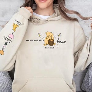 May include: A beige hoodie with a Winnie the Pooh inspired design. The front of the hoodie says "mama bear est. 2025" with a cartoon Winnie the Pooh holding a honey pot. The left sleeve has a cartoon Winnie the Pooh holding a balloon with the text "I love you" and a heart.