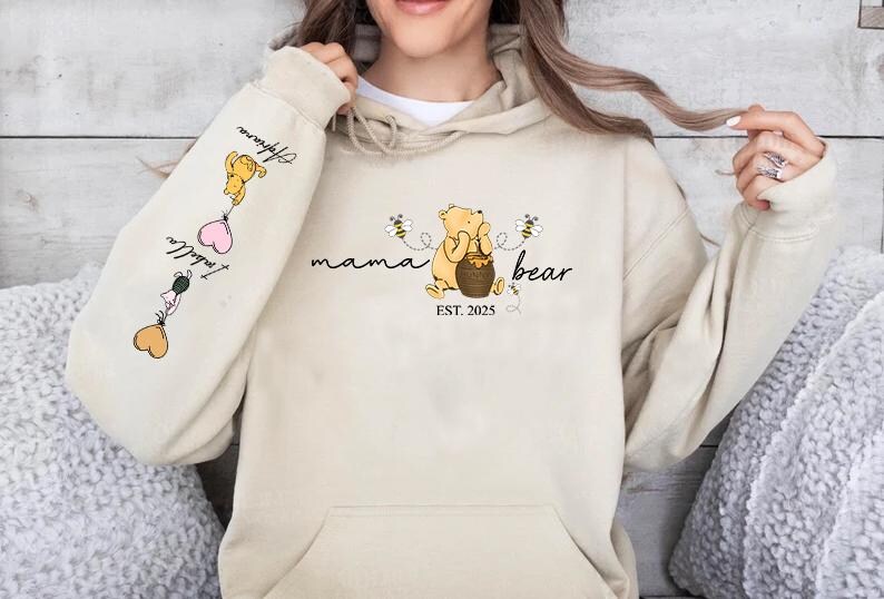 May include: A beige hoodie with a Winnie the Pooh inspired design. The front of the hoodie says "mama bear est. 2025" with a cartoon Winnie the Pooh holding a honey pot. The left sleeve has a cartoon Winnie the Pooh holding a balloon with the text "I love you" and a heart.