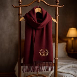 May include: A burgundy scarf with a fringed edge hangs on a wooden coat rack. The scarf has an embroidered gold floral design with the name "Lily". The scarf is made of a soft material.