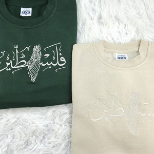 May include: Two sweatshirts with a white embroidered design of a map of Palestine and the Arabic text "Palestine" on a green and beige sweatshirt.