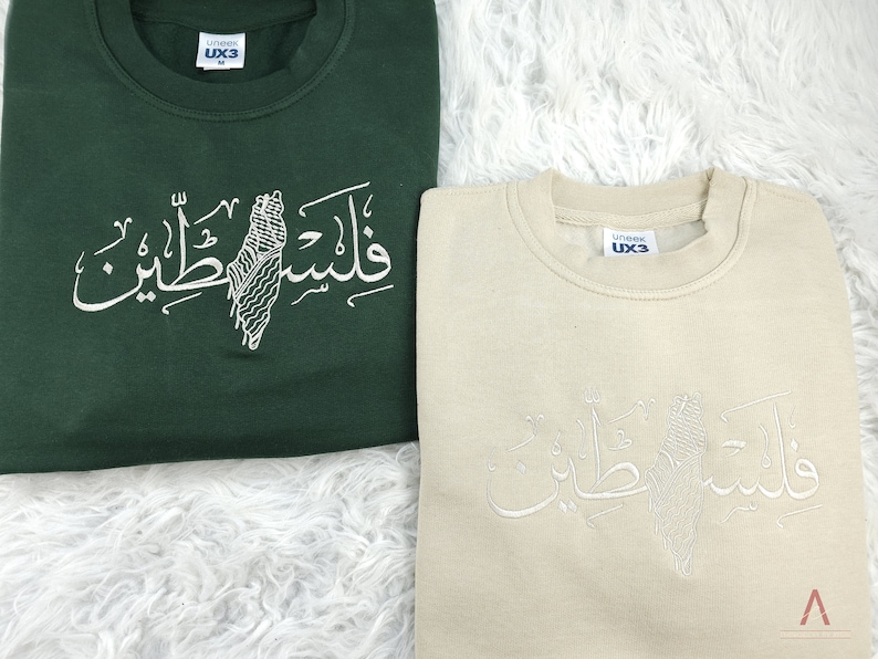 May include: Two sweatshirts with a white embroidered design of a map of Palestine and the Arabic text "Palestine" on a green and beige sweatshirt.
