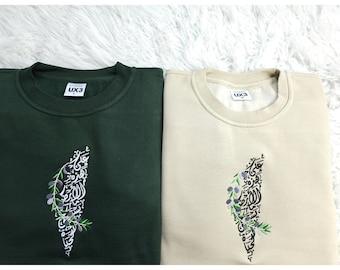 May include: Two sweatshirts, one green and one beige, with an embroidered design of an olive branch and Arabic calligraphy in the shape of the country of Palestine. The sweatshirts have a label with the text "UX3" on them.