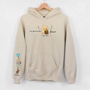 May include: A beige hoodie with a Winnie the Pooh inspired design. The front of the hoodie says "mama bear est. 2025" with a cartoon Winnie the Pooh holding a honey pot. The left sleeve has a vertical design of Winnie the Pooh characters with balloons.
