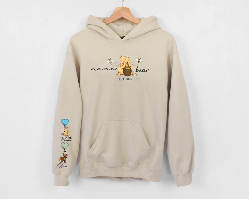 May include: A beige hoodie with a Winnie the Pooh inspired design. The front of the hoodie says "mama bear est. 2025" with a cartoon Winnie the Pooh holding a honey pot. The left sleeve has a vertical design of Winnie the Pooh characters with balloons.