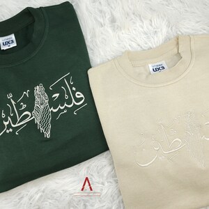 May include: Two sweatshirts with Arabic calligraphy and a stylized image of Palestine. The sweatshirt on the left is green and the sweatshirt on the right is beige.