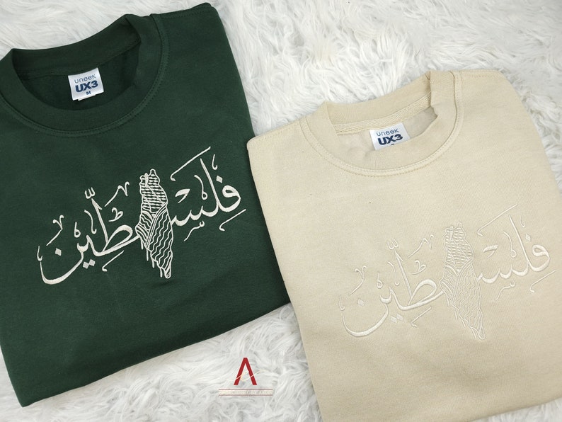 May include: Two sweatshirts with Arabic calligraphy and a stylized image of Palestine. The sweatshirt on the left is green and the sweatshirt on the right is beige.