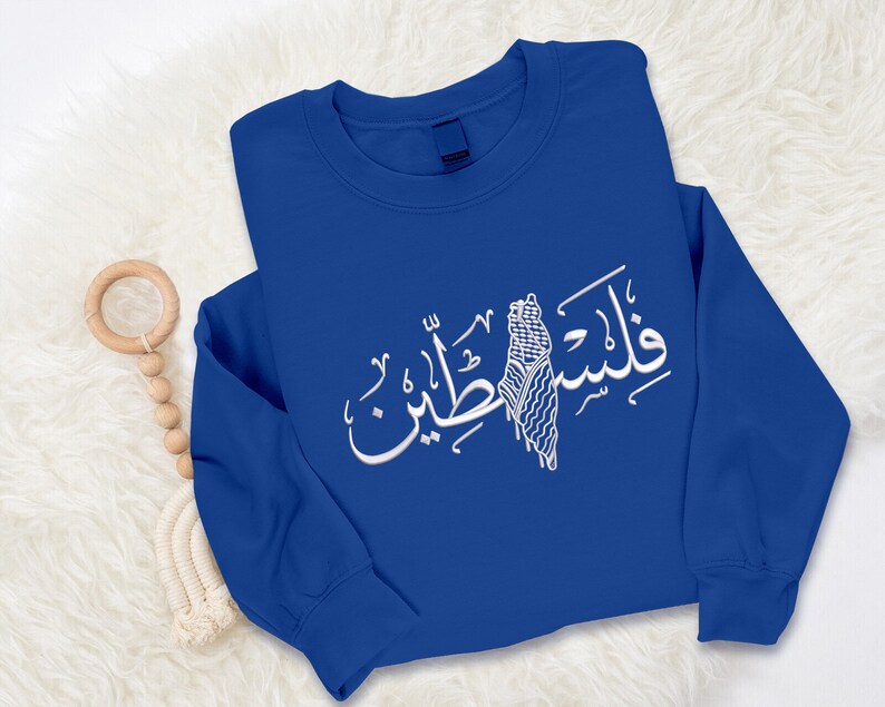 May include: A royal blue sweatshirt with a white Arabic calligraphy design featuring the word "Palestine" and a stylized image of the Palestinian flag.