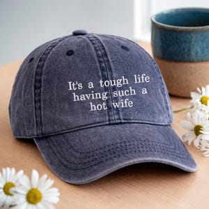 May include: A dark blue, washed-out cotton baseball cap featuring white embroidered text on the front that reads, "It's a tough life having such a hot wife." The cap has a curved brim, a button on top, and visible stitching along the panels.