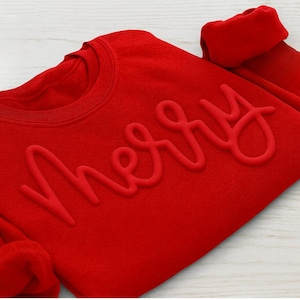 Puede incluir: Una sudadera roja de cuello redondo con la palabra "Merry" en relieve, en fuente cursiva. Las mangas están dobladas. La sudadera está hecha de un material suave, ideal para la temporada navideña.