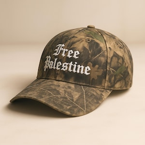 May include: A camouflage baseball cap with the words "Free Palestine" embroidered in white script across the front. The cap features a curved brim and a button on top. The camouflage pattern includes shades of brown, green, and tan.