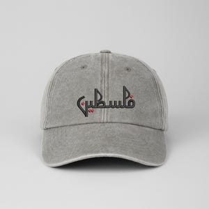 May include: Grey baseball cap featuring the Arabic word "Palestine" embroidered in black and red. The cap has a curved peak and an adjustable strap. The hat is made from a soft, washed cotton material.