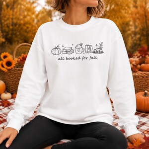 Puede incluir: Una sudadera blanca de manga larga con cuello redondo que presenta un gráfico de líneas negras de una calabaza, libros apilados, una taza con un corazón y una bufanda a cuadros. Debajo, el texto all booked for fall en letra cursiva.