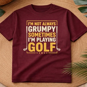 May include: Maroon t-shirt with the text "I'M NOT ALWAYS GRUMPY SOMETIMES I'M PLAYING GOLF" in white and yellow. The design includes golf clubs and stars. The shirt is displayed on a woven surface.