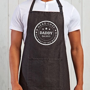 May include: A dark grey apron with a large front pocket and adjustable neck strap. The apron features a white circular design with the words "HEAD CHEF DADDY Est. 2015 THE BEST IN THE WORLD".