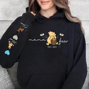 May include: Black hoodie with a Winnie the Pooh illustration and the text "mama bear est. 2025". The sleeve has a series of hearts and illustrations of Winnie the Pooh characters.
