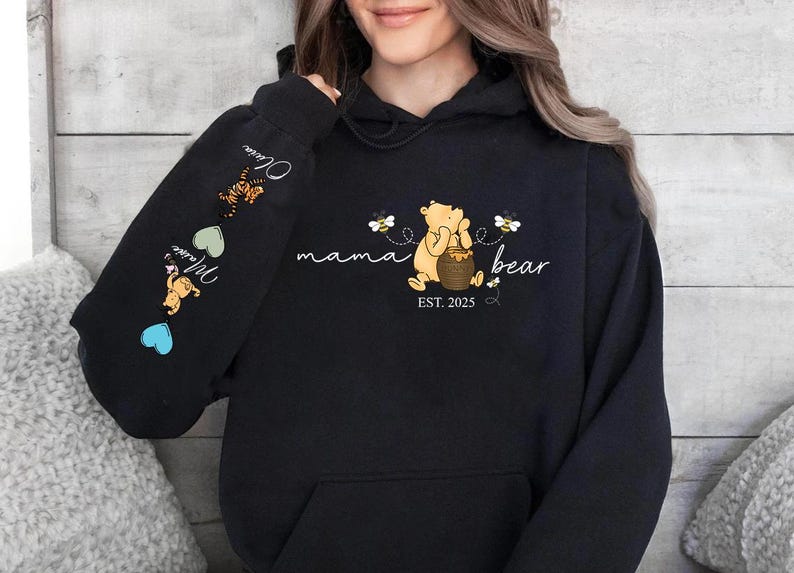 May include: Black hoodie with a Winnie the Pooh illustration and the text "mama bear est. 2025". The sleeve has a series of hearts and illustrations of Winnie the Pooh characters.