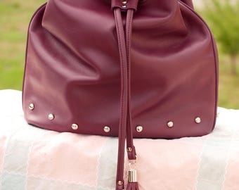 May include: A burgundy leather bucket bag with a drawstring closure and silver metal accents. The bag has a tassel on the bottom.