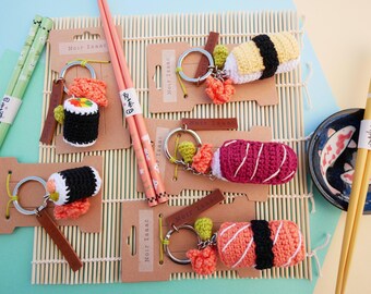 May include: A collection of handmade sushi keychains, including nigiri and maki rolls, displayed on kraft paper tags. Chopsticks in various colors and a small dish with koi fish are also visible. The keychains have metal rings and leather tags.