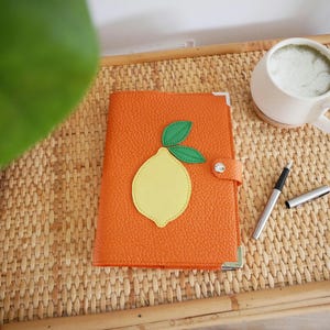 May include: Orange leather journal with a lemon applique. The journal is closed with a snap closure. A white mug with a green beverage and a silver pen are on a woven tray.