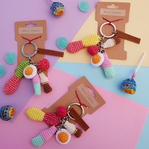 May include: Several handmade crochet keyrings in vibrant colours. Each keyring has a unique design with red, yellow, green, and white elements. The keyrings are attached to a silver ring with a brown leather tag that reads "Noir Isaac".