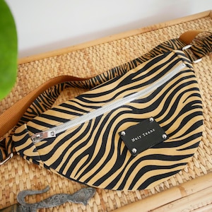Peut inclure: Sac banane à rayures zébrées avec fermeture éclair argentée et étiquette en cuir noir « Noir Isaac ». Le sac a une sangle marron et est posé sur une surface tissée.