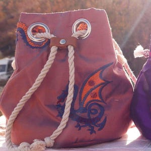 May include: A burgundy drawstring bag with a dragon design embroidered in orange and blue. The bag has a rope drawstring closure and metal grommets. The dragon has orange wings and is set against a burgundy background. The bag is made of a soft fabric.