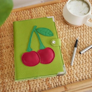 May include: A green leather journal with a cherry design. The journal has a red cherry applique with green stems and leaves. A silver pen and a white mug with a beverage are on a woven tray.