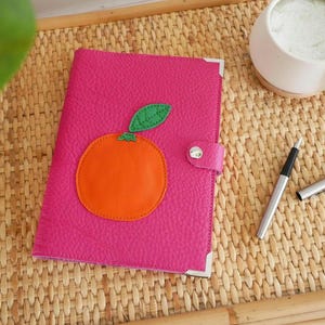 May include: A fuchsia leather journal with an orange and green applique of an orange. The journal has silver corner protectors and a snap closure. A silver pen and a cup of matcha are also on the woven surface.
