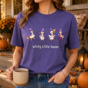 May include: A purple t-shirt featuring a graphic of four white geese wearing purple witch hats and capes, with the text "Witchy Little Goose" printed below. The geese are depicted in various poses, including holding a broom, a wand, and a cauldron.