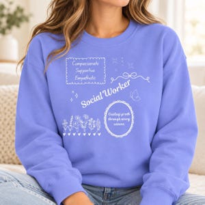 May include: A periwinkle blue crewneck sweatshirt featuring white line art graphics and text. The design includes the words "Compassionate Supportive Empathetic", "Social Worker", and "Guiding growth through every season", alongside floral, butterfly, and star motifs.