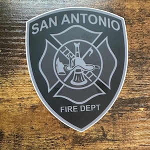 May include: A shield-shaped sticker with the words "SAN ANTONIO" at the top and "FIRE DEPT" at the bottom. The center features a fire department emblem in gray and white.